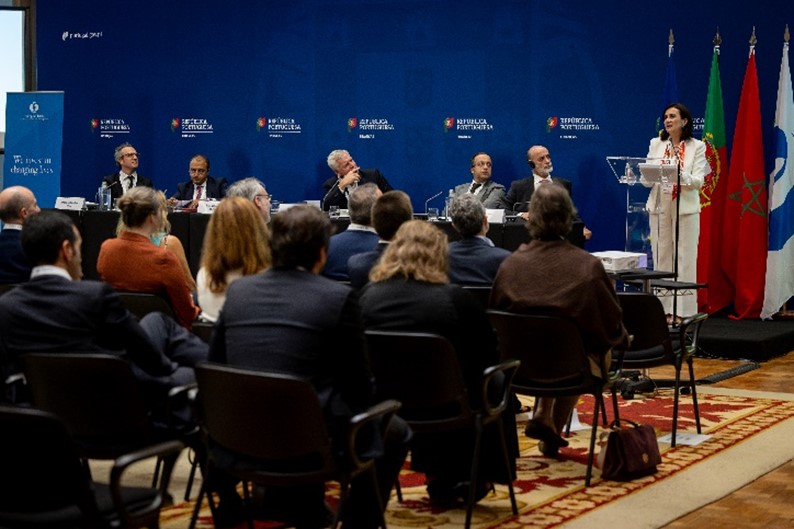 Salão Nobre do Ministério das Finanças com plateia e mesa dos oradores. Pessoa a discursar.