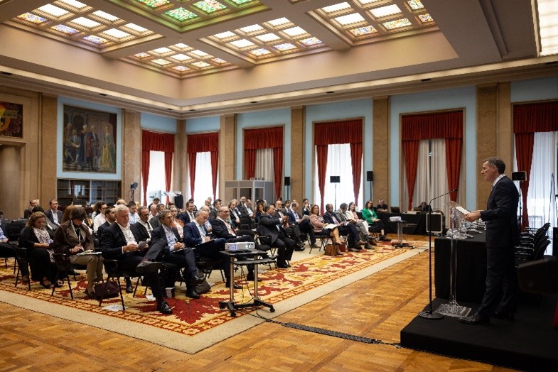 Salão Nobre do Ministério das Finanças com plateia e pessoa a discursar. Visualiza-se o teto da sala com vitrais, pintura ao fundo e janelas com pendões vermelhos.