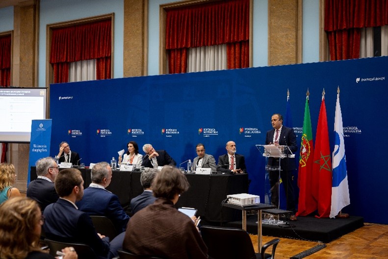Salão Nobre do Ministério das Finanças com plateia e mesa dos oradores. Pessoa em pé no púlpito a discursar com bandeiras atrás (UE, Portugal, Marrocos e BERD).