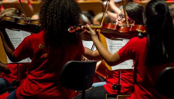 Três raparigas da Orquestra Geração sentadas a tocar violino. A farda da orquestra é uma t-shirt vermelha. Pode-se visualizar também as respetivas pautas e outros elementos ao fundo.