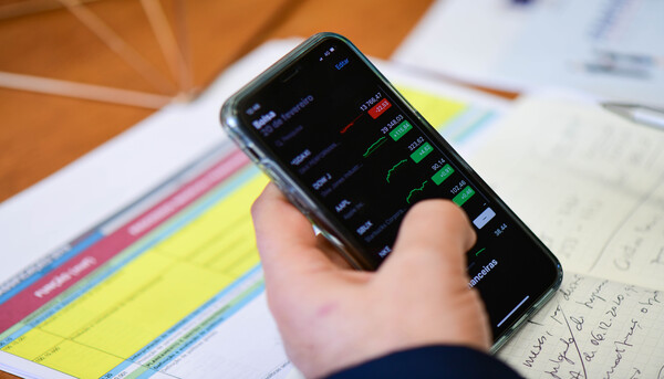 Mão com telemóvel em cima de papeis e caderno. No ecrã no telemóvel visualiza-se cotações de bolsa.