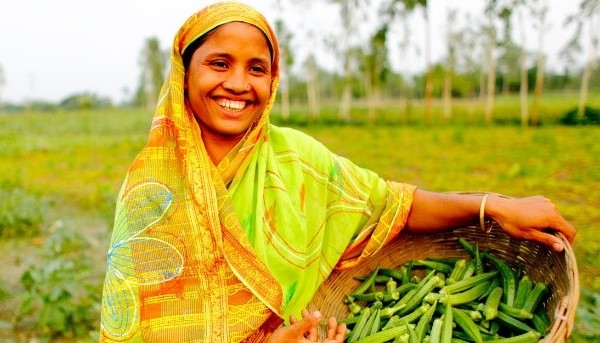 Mulher a sorrir num campo agrícola segurando uma cesta de quiabos.