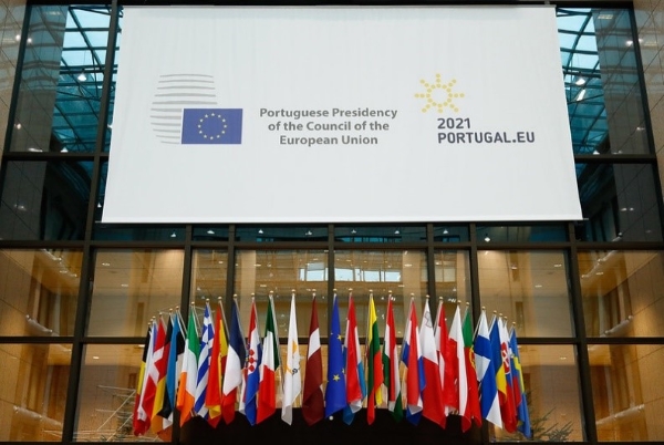 Edifício envidraçado com as bandeiras da união europeia. Por cima das bandeiras está um flyer com o logo da UE, o descritivo 