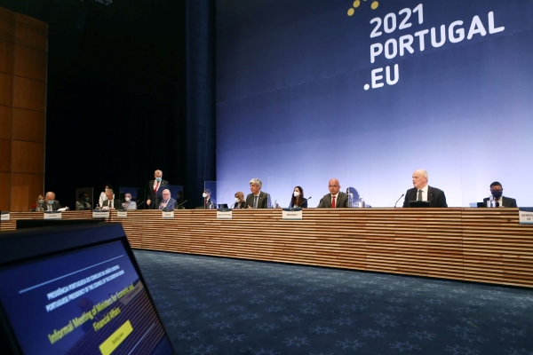 Pode visualizar-se mesa com os ministros das finanças sentados lado a lado. Como fundo está projetado uma imagem azul com o logo da presidência europeia portuguesa e com a descrição 2021.PORTUGAL.EU.