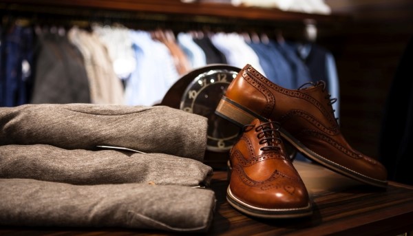 Na imagem visualiza-se um par de sapatos castanhos e camisolas de homem ao centro. Atrás vários cabides de roupa em exposição.