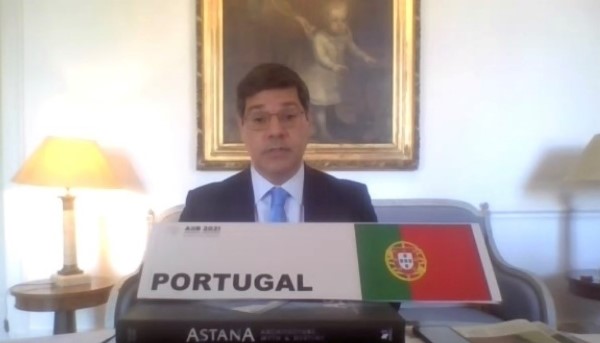 Imagem de representante de Portugal a discursar na sua secretária. Há frente da secretária pode-se observar a bandeira portuguesa e atrás um candeeiro do lado esquerdo com um sofá ao centro e um quadro por cima.