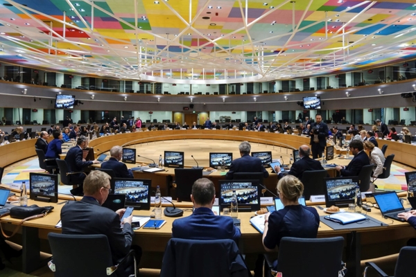 Sala do conselho europeu com pessoas sentadas na mesa circular em madeira com computadores integrados a toda a volta. A sala é composta por televisores em cima a toda a volta e o teto tem estrutura aos quadrados em diversas cores (tons de vermelho, azul, verde, amarelo, roxo entre outras).