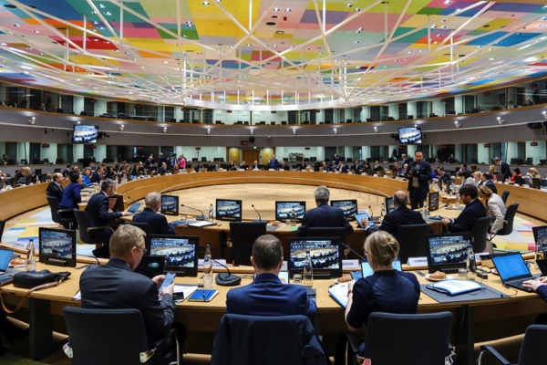 Sala do conselho europeu com pessoas sentadas na mesa circular em madeira com computadores integrados a toda a volta. A sala é composta por televisores em cima a toda a volta e o teto tem estrutura aos quadrados em diversas cores (tons de vermelho, azul, verde, amarelo, roxo entre outras). 