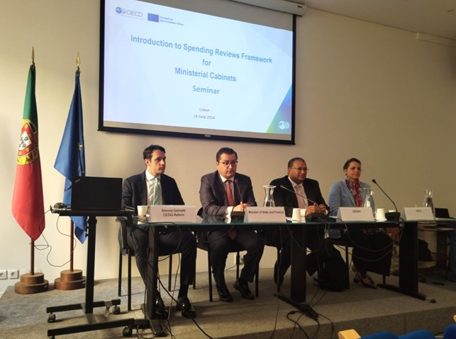 Auditório - Oradores na mesa (ao centro: Ministro das Finanças, Joaquim Miranda Sarmento e Diretor-Geral do GPEARI, José Carlos Pereira) com bandeiras Portugal e UE ao fundo. Slide projetando emblema da UE e logotipo da OCDE.